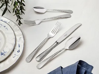 A Villeroy & Boch Leandra cutlery set elegantly arranged on a white table. It consists of two forks, two spoons and a knife. A plate with a blue rim and a blue napkin are partially visible, while green foliage adds a charming decorative touch to the table.