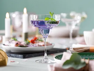 Ein violetter Cocktail, garniert mit Minze, in einem Purismo Margarita Glas auf einem gedeckten Tisch, umgeben von Tellern, Kerzen und Dekorationsgegenständen.