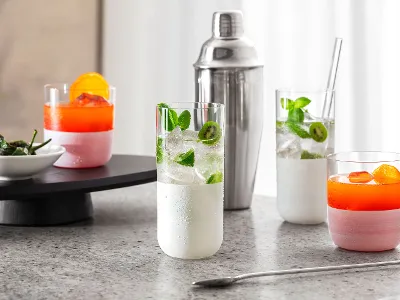 Vier bunte Cocktails in weißen Manufacture Gläsern auf einer Marmoroberfläche, im Hintergrund ein Cocktailshaker und auf einem Tablett eine kleine Schale mit Paprika.