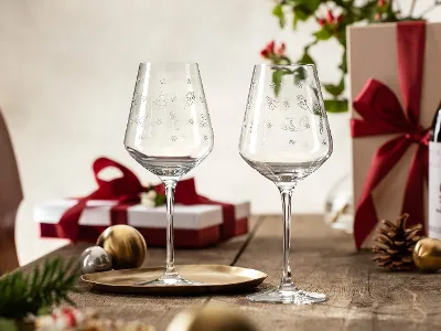 Ein Paar Toy’s Delight-Weingläser von Villeroy & Boch stehen auf einem Tisch inmitten verpackter Geschenke, einer Weinflasche und festlicher Dekorationen.