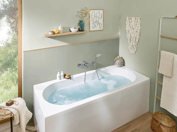 Mit Wasser gefüllte O.novo-Badewanne Ein modernes Badezimmer mit einer mit Wasser gefüllten Badewanne der Kollektion O.novo, einem Wandregal mit Toilettenartikeln, einem Makramee-Wandbehang und einem Fenster mit Blick ins Grüne.