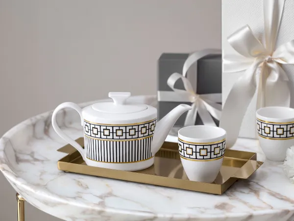 A white-and-gold patterned MetroChic teapot and cups on a gold tray on a marble table. Two wrapped presents and a small white flower are visible in the background.