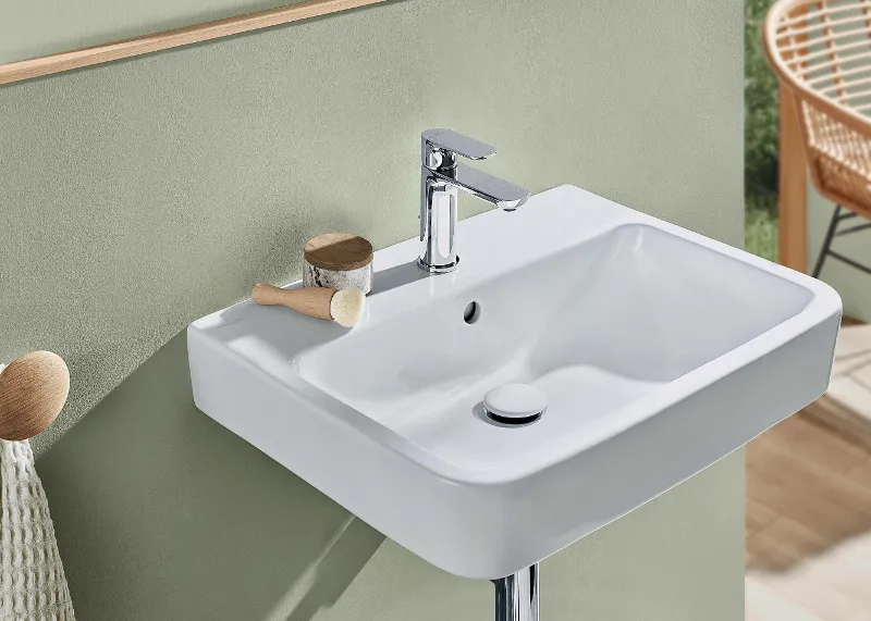 Weißes O.novo-Waschbecken mit verchromter Armatur Ein modernes rechteckiges weißes O.novo-Waschbecken mit verchromtem Wasserhahn, Bürste und kleinem Behälter an der Rückwand vor einer mattgrünen Wand. Im Hintergrund steht ein beiger Stuhl.