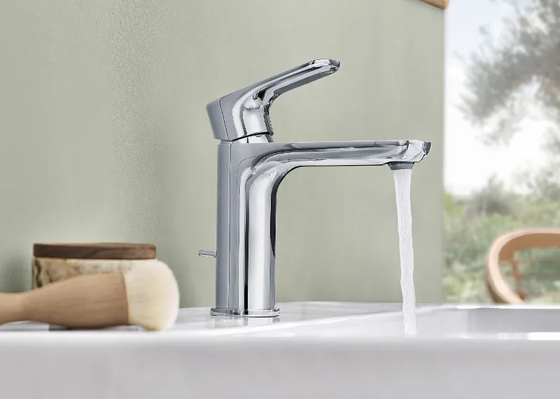 Moderne Chromarmatur mit fließendem Wasser Auf einem weißen O.novo-Waschbecken ist eine moderne Chromarmatur mit fließendem Wasser installiert. Daneben stehen eine Bürste und ein Holzbehälter.