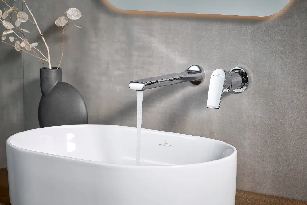 Ein modernes Waschbecken im Badezimmer, aus dessen verchromter Armatur Wasser fließt. Neben dem Villeroy & Boch Collaro Waschbecken steht eine schwarze Vase mit getrockneten Pflanzen.
