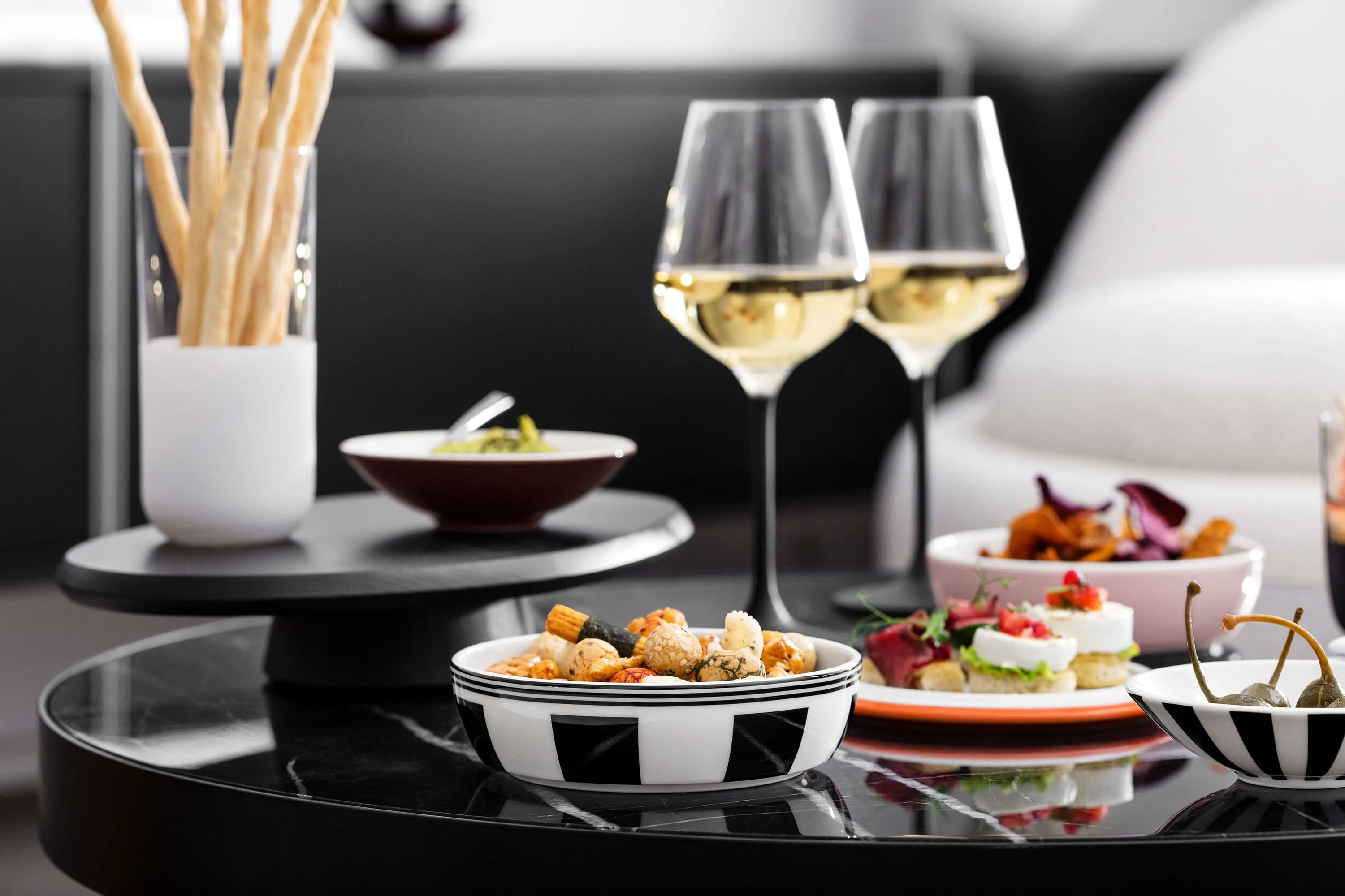 La petite Boule pleine d’amuse-bouches et de verres à vin blanc Manufacture. Une table noire avec des coupelles La petite Boule remplies d’amuse-bouches et de verres à vin blanc Manufacture.