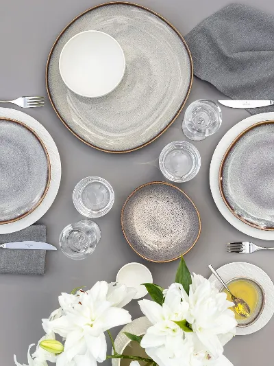 Vista superior de una mesa de comedor con dos vajillas, compuestas por platos Lave beige y platos Manufacture blancos, cubiertos, vasos y un jarrón con flores blancas.