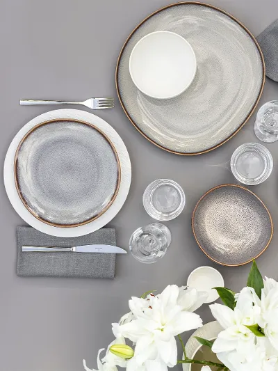 Vista superior de una mesa de comedor con dos vajillas, compuestas por platos Lave beige y platos Manufacture blancos, cubiertos, vasos y un jarrón con flores blancas.