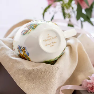 Une tasse de l'Annual Easter Edition est posée à l'envers sur un tissu beige et montre le cachet de l'année. Une tasse de l'Annual Easter Edition de Villeroy & Boch est posée à l'envers sur un tissu beige et montre son cachet annuel doré, tandis qu'une fleur rose et un vase avec des feuilles sont visibles en arrière-plan.