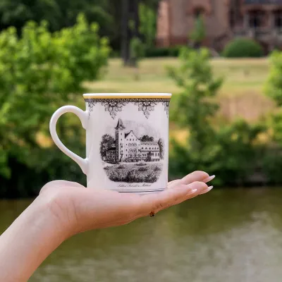 Audun cup in front of a river and trees A hand holding a decorative Audun cup with an illustration of a building in front of a river and trees.
