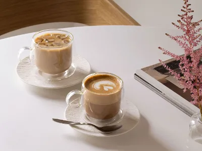 Zwei Villeroy & Boch Artesano-Glasbecher auf einem Tisch Zwei Villeroy & Boch Artesano-Glasbecher auf einem Tisch: einer mit Eiskaffee gefüllt, der andere mit Latte, dazu ein Löffel, ein Stapel Zeitschriften und eine rosa Blume in einer Vase.