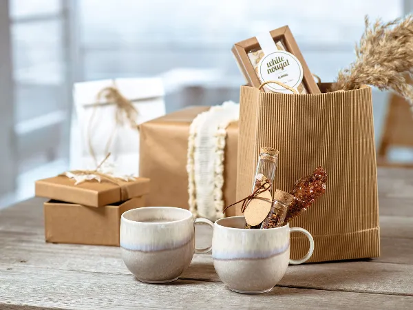 Perlemor Kaffeebecher vor braunen Geschenkboxen aus Papier Braune Geschenkboxen mit rustikaler Ästhetik, davor von zwei Keramikbechern aus der Kollektion Perlemor von Villeroy & Boch auf einem Holztisch.