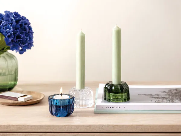 Auf einem Holztisch steht eine Fleur Vase und Fleur Teelichthalter von Villeroy & Boch ein blaues Blumenarrangement schmückt die Vase Auf einem Holztisch steht eine Fleur Vase und Fleur Teelichthalter von Villeroy & Boch ein blaues Blumenarrangement schmückt die Vase, eine brennende Kerze in einem blauen Halter, zwei grüne Kerzen in Haltern, ein Buch und Streichhölzer.