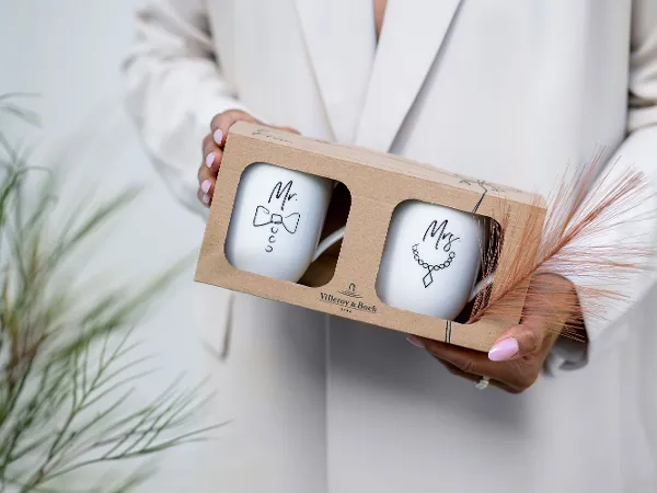 Eine Person hält einen Geschenkkarton mit zwei Statement Tassen in der Hand. Eine Person hält einen Geschenkkarton, der zwei Statement Tassen mit der Aufschrift „Mr.“ und „Mrs.“ enthält in der Hand.