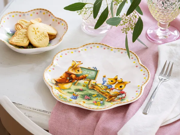 Les assiettes décoratives de l'édition Annual Easter de Villeroy & Boch avec des lapins. Les assiettes décoratives de l'édition Annual Easter de Villeroy & Boch avec des lapins sont élégamment disposées sur une table avec une serviette rose, une fourchette et un bol de biscuits.