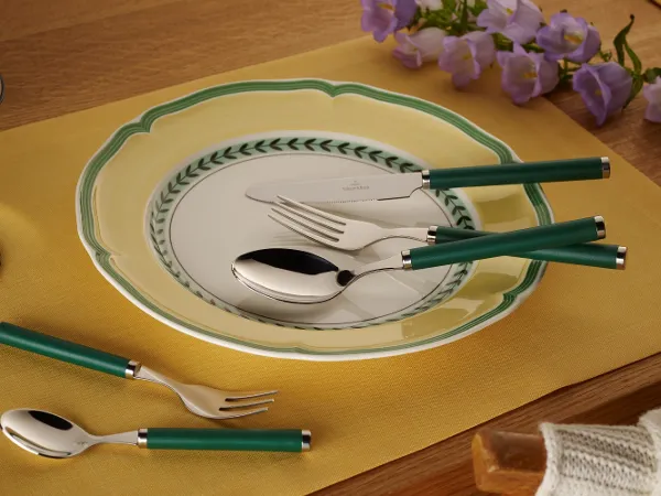 A dining room set with the Villeroy & Boch Play! collection, featuring a decorative plate, cutlery with green handles and a yellow placemat on a wooden table with purple flowers in the background.