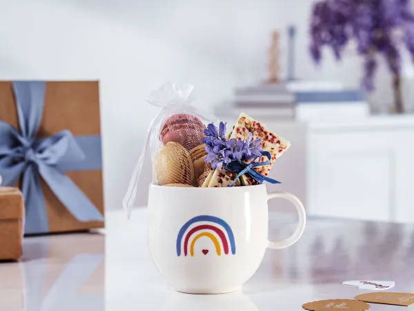 Eine „With Love“-Tasse von Villeroy & Boch mit Regenbogendesign gefüllt mit Macarons Eine „With Love“-Tasse von Villeroy & Boch mit Regenbogendesign, gefüllt mit Macarons, einer Tafel Schokolade und lila Blumen auf einer weißen Oberfläche mit Geschenkboxen und Büchern im Hintergrund.