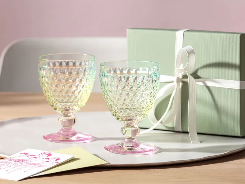 Zwei Boston-Gläser von Villeroy & Boch mit rosafarbenem Boden stehen auf einem Tablett neben einer grünen Geschenkbox mit weißer Schleife und einer Grußkarte auf einem Holztisch.