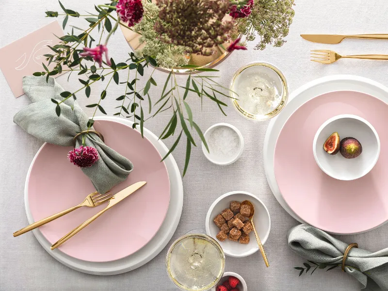 Gedeckter Tisch mit Rose Garden Tellern und goldenem MetroChic Besteck Elegante Tischdekoration mit rosa Rose Garden Tellern, goldenem MetroChic Besteck, frischen Blumen und einem Hauch von Grün, dazu Getränke und leichte Snacks.