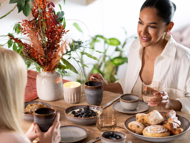 Two women sitting opposite each other at a table set with pastries, berries and coffee. One woman has her hair tied back and they are chatting. There is a vase of flowers on the table.
