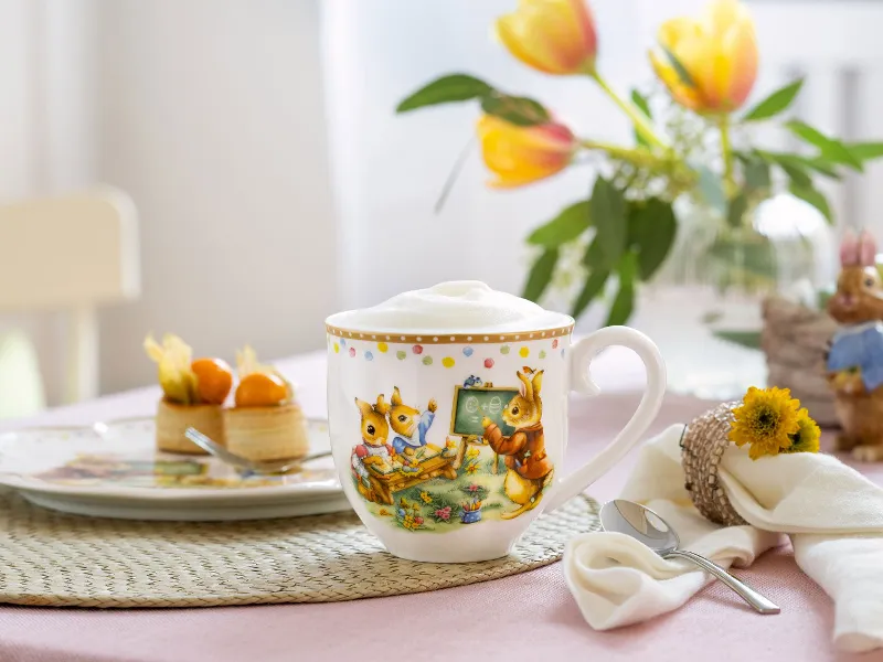Tasse de l'Annual Easter Edition de Villeroy & Boch décorée d'un motif de lapin Le gobelet décoratif avec couronne de mousse de l'Annual Easter Edition de Villeroy & Boch, décoré d'un motif de lapin, est posé sur une table à côté d'une assiette à dessert, d'une nappe fleurie et d'une figurine de lapin. Des tulipes sont en fleurs à l'arrière-plan.