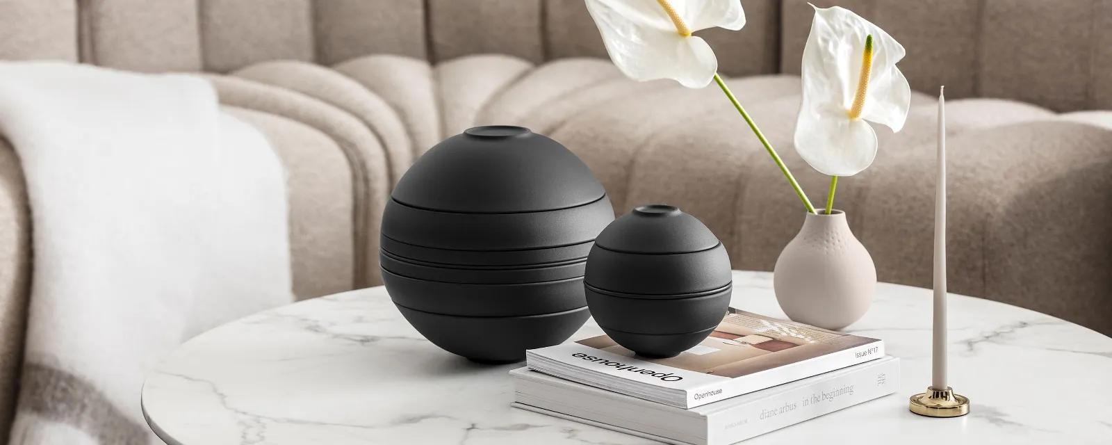 Die schwarze La Boule von Villeroy & Boch in einem beigen Wohnzimmerambiente. Die schwarze La Boule von Villeroy & Boch, einer beigen Vase mit weißen Blumen, einem Kerzenständer und einigen Büchern in einem beigen Wohnzimmerambiente.