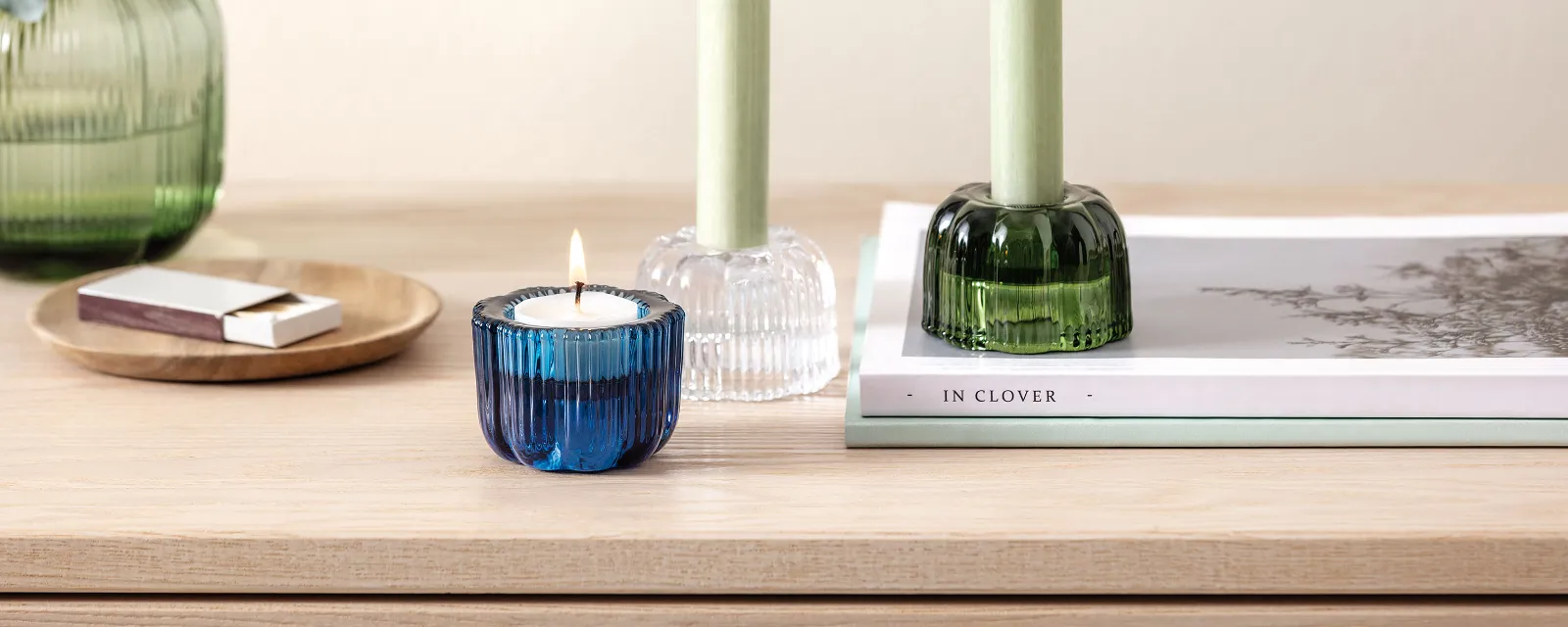 Auf einem Holztisch steht eine Fleur Vase und Fleur Teelichthalter von Villeroy & Boch ein blaues Blumenarrangement schmückt die Vase Auf einem Holztisch steht eine Fleur Vase und Fleur Teelichthalter von Villeroy & Boch ein blaues Blumenarrangement schmückt die Vase, eine brennende Kerze in einem blauen Halter, zwei grüne Kerzen in Haltern, ein Buch und Streichhölzer.