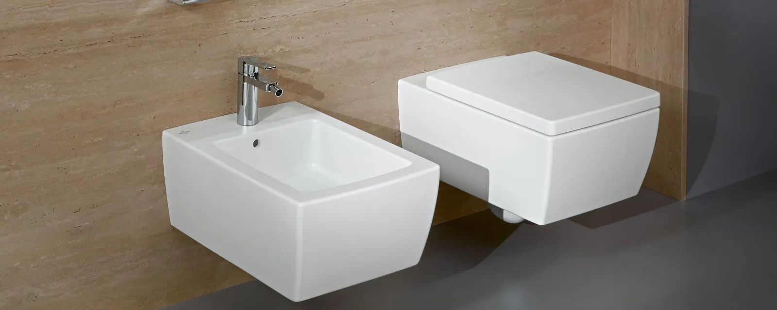 Weiße Toilette und Bidet aus der Kollektion Memento 2.0 Modernes Bad mit weißem Wand-WC und Bidet vor beige gefliesten Wänden, ausgestattet mit verchromter Armatur, Toilettenspülung und elegantem Handtuchhalter.