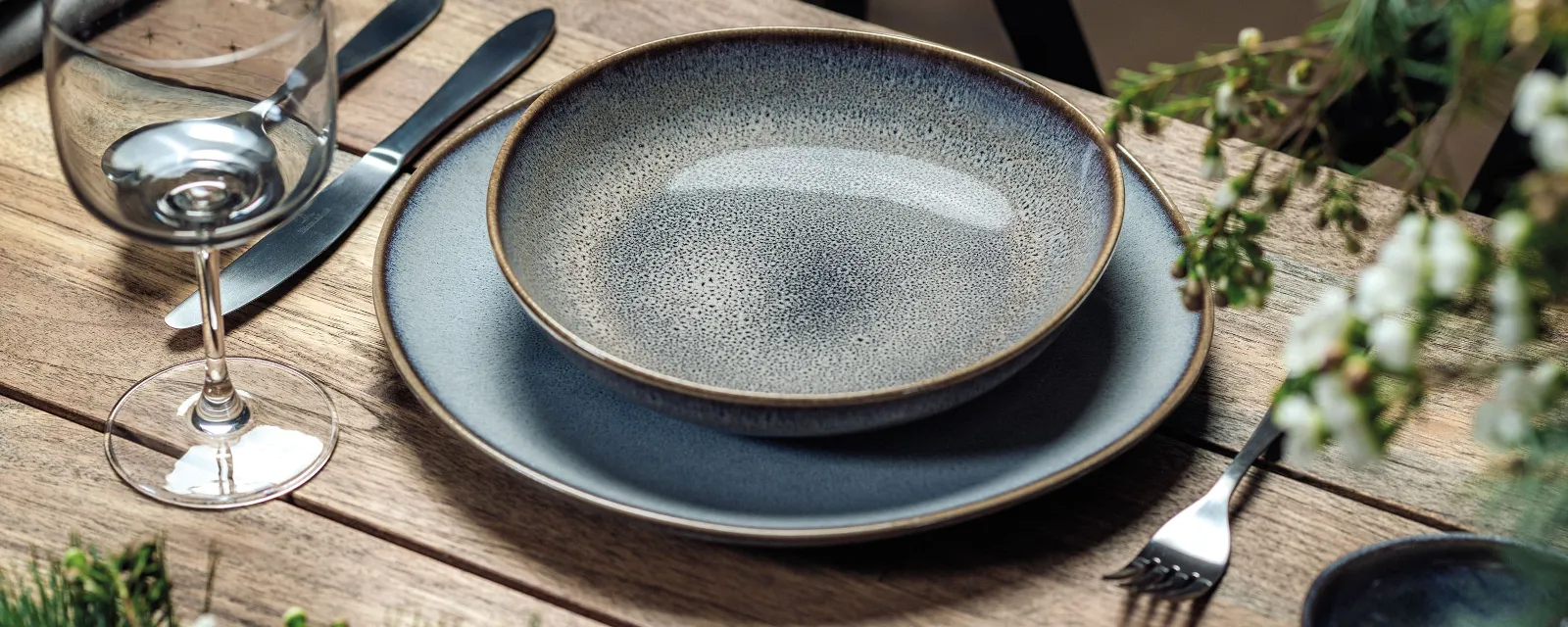Stilvolle Esstischgarnitur mit Lave Tellern und Besteck auf Holztisch Elegante Esstischgarnitur mit Lave Tellern, Besteck und einem Glas auf einem Holztisch mit grünem Laub.