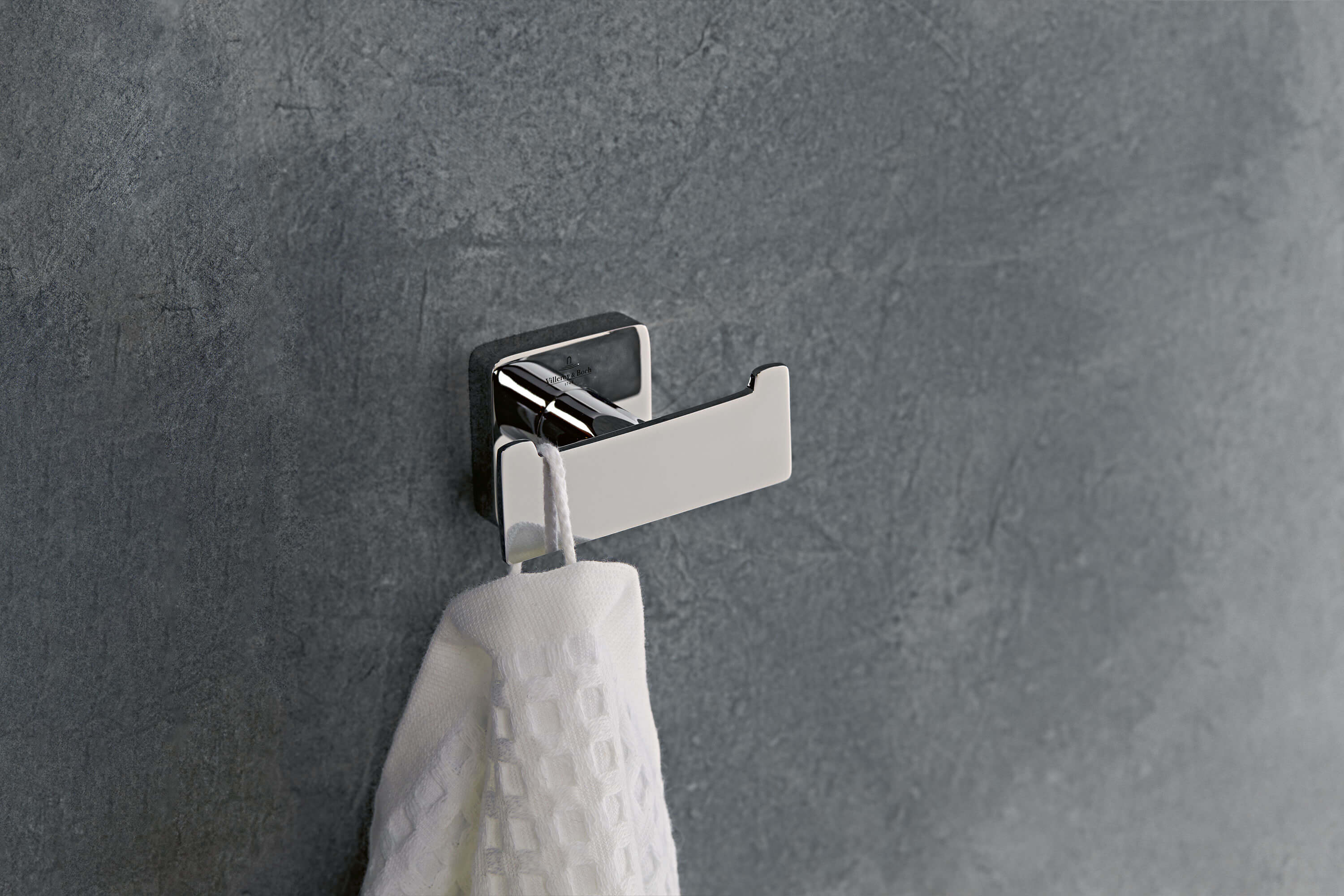 Ein weißes Handtuch hängt an einem modernen, metallischen Wandhaken vor einer grau strukturierten Wand.